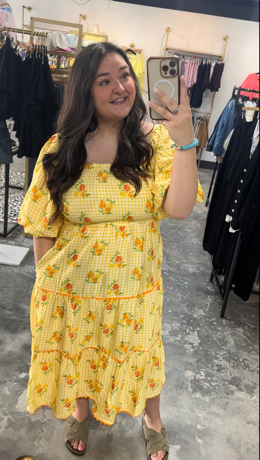 Woman taking a mirror selfie wearing a yellow dress with floral patterns in a clothing store.