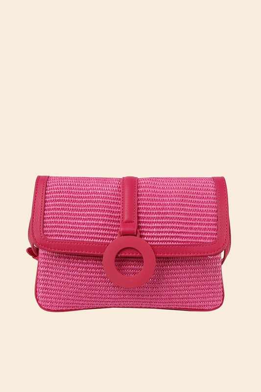 A pink straw crossbody bag with a flap closure and a ring detail on the front.