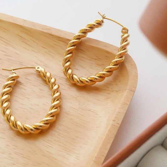 A pair of gold-colored braided hoop earrings displayed on a wooden surface.