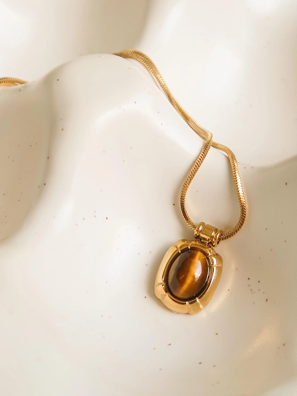 A gold-colored necklace with a cat-eye stone pendant, displayed on a white ceramic dish.